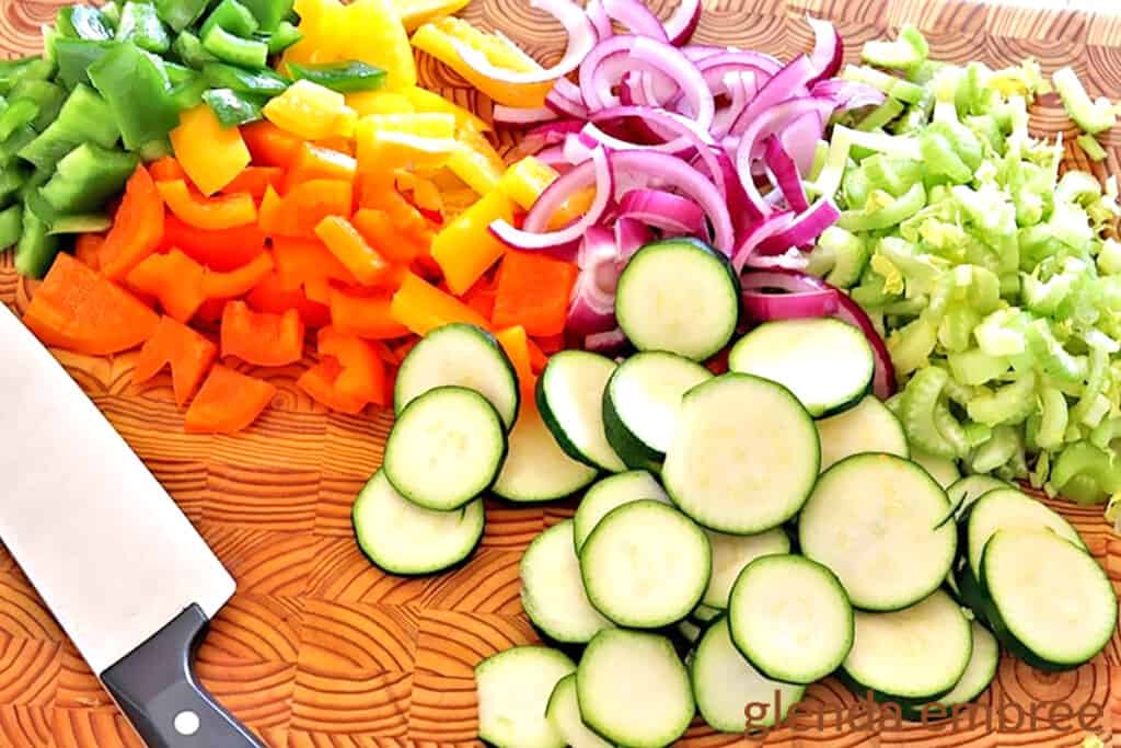 sliced veggies on a wooden cutting board with a chef's knife: zucchini slices, celery slices, slivered red onion,s red, yellow and green bell peppers