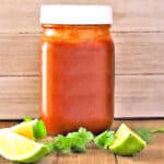 Homemade Enchilada Sauce in a glass mason jar on a wooden table with lime segments and sprigs of fresh cilantro
