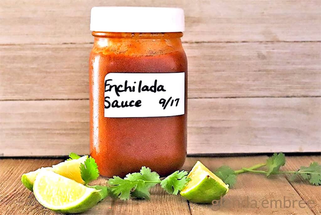 Homemade Enchilada Sauce in a glass mason jar on a wooden table with lime segments and sprigs of fresh cilantro