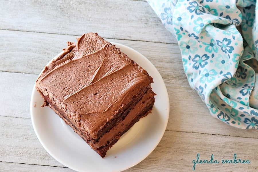 homemade chocolate cake with fudge frosting on a white plate. Plate is sitting on a wooden table and there is a blue and white gingham napkin on the right.