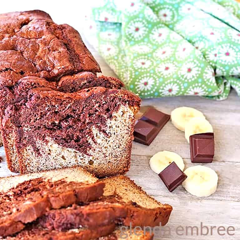 chocolate swirl banana bread on a rough wooden table with squares of baking chocolate and banana slices next to a green print fabric napkin