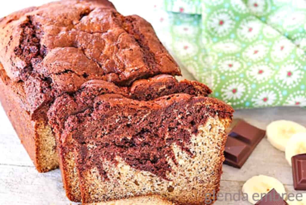 chocolate swirl banana bread on a rough wooden table with squares of baking chocolate and banana slices next to a green print fabric napkin