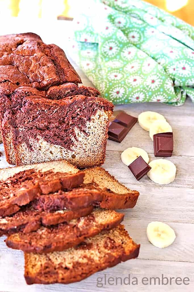 chocolate swirl banana bread on a rough wooden table with squares of baking chocolate and banana slices next to a green print fabric napkin