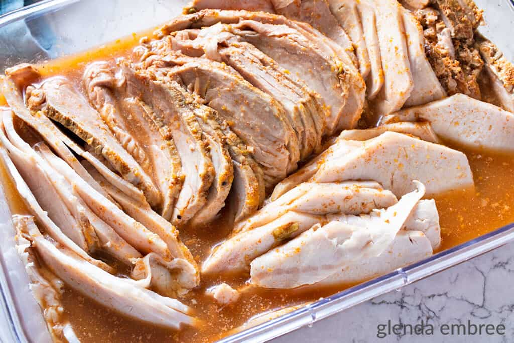 Slow Cooker Pork carnitas roast, sliced thin and stored in its cooking liquid in a clear rectangular container with no lid.