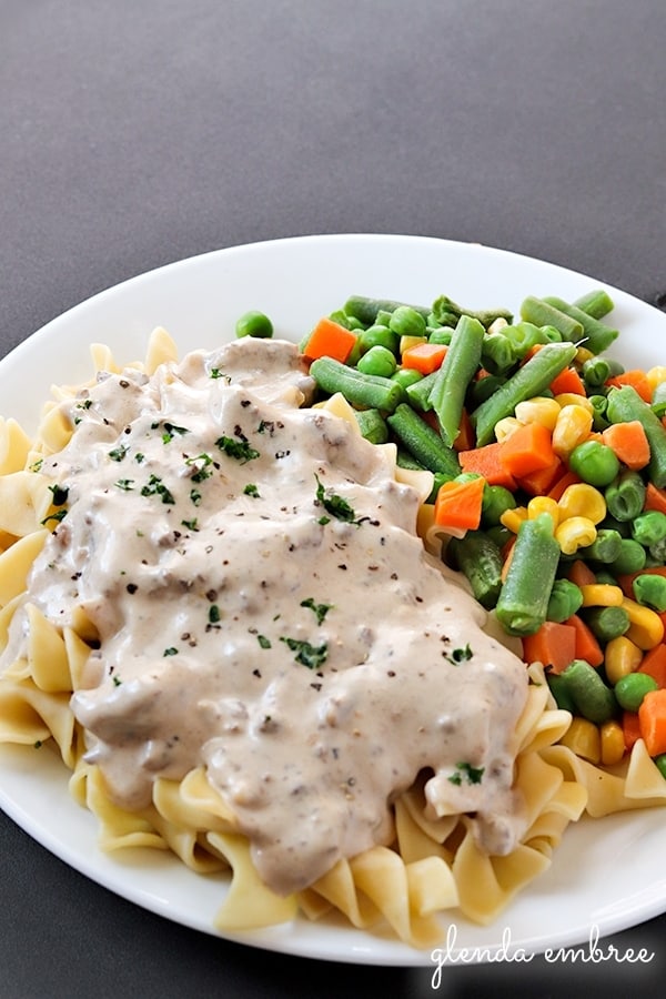 Hamburger Stroganoff over egg noodles served on a white plate with mixed vegetables.