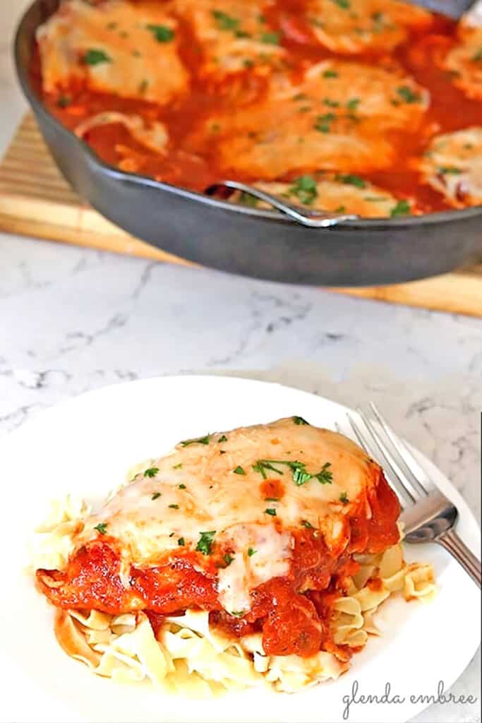 Chicken Parmesan on a bed of noodles on a white plate. Cast iron skillet of chicken parmesan in the background.