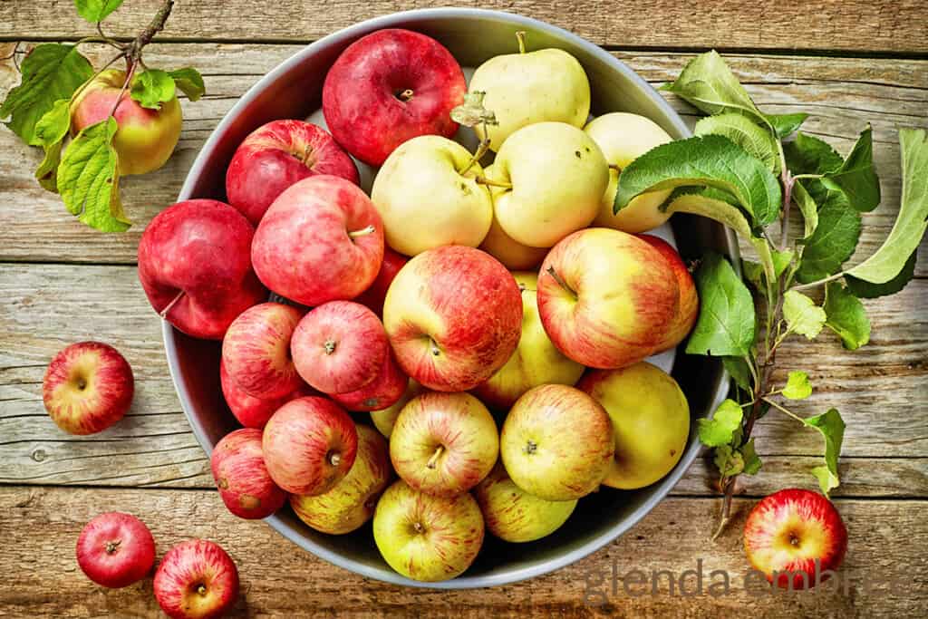 red and yellow apples in a stainless steel bowl.  Bowl is resting on a rough hewn wooden work bench