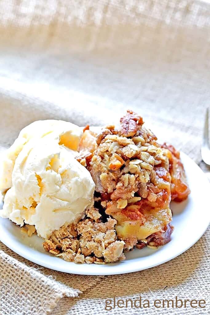 homemade apple crisp with a scoop of vanilla ice cream on a white plate.  Plate is sitting on brown burlap fabric