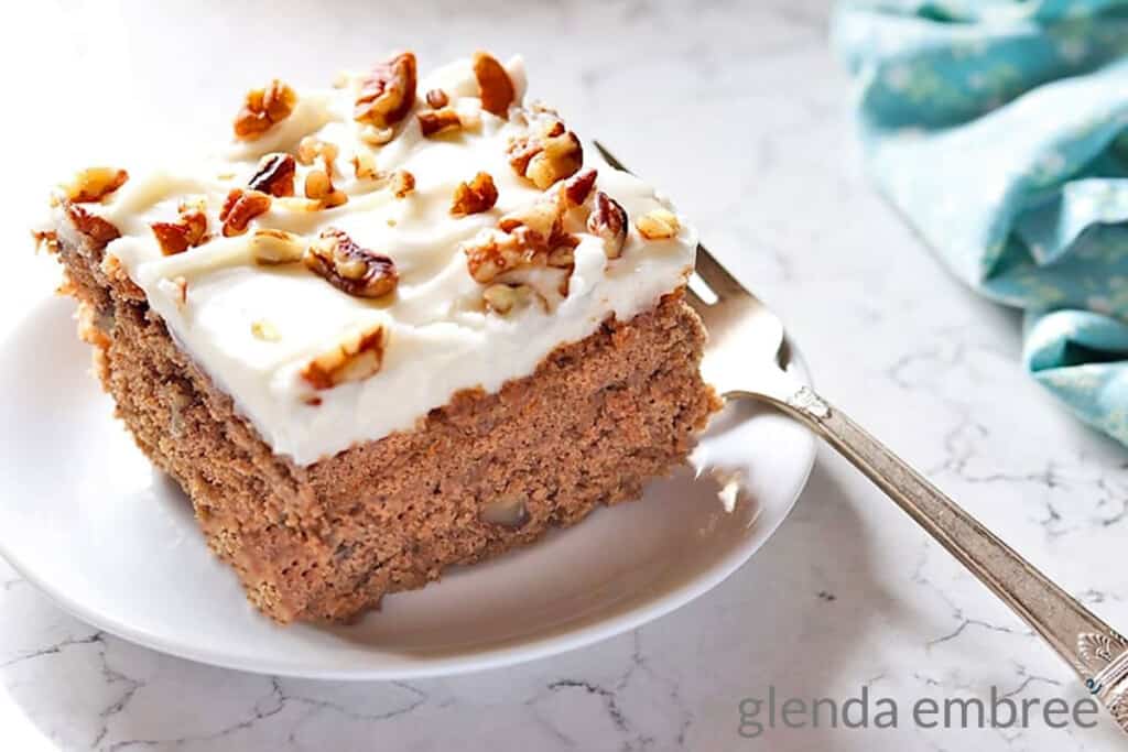 Sour Cream Spice Cake on a white plate. Plate is on a marble countertop and has a fork and blue print fabric napkin next to it. Cake is frosted with cream cheese icing and sprinkled with chopped pecans.