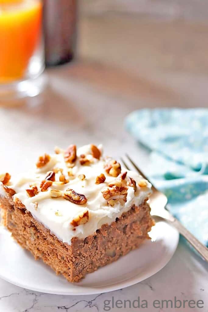 Sour Cream Spice Cake on a white plate.  Plate is on a marble countertop and has a fork and blue print fabric napkin next to it.  Cake is frosted with cream cheese icing and sprinkled with chopped pecans.