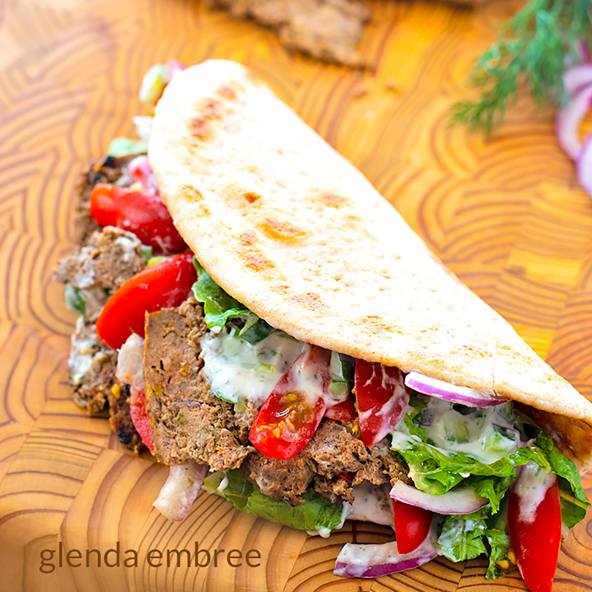 homemade gyro meat in a pita flatbread with lettuce, tomatoes, red onion and tzatziki on a wooden cutting board