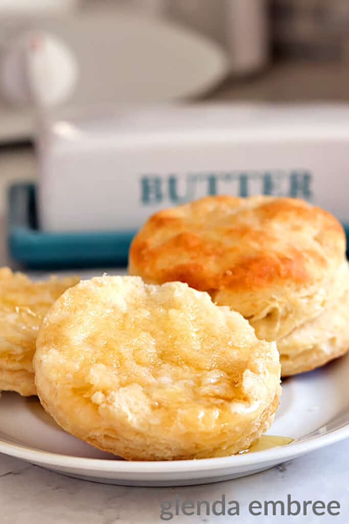 homemade biscuits on a white plate slathered with creamy butter and dripping with honey
