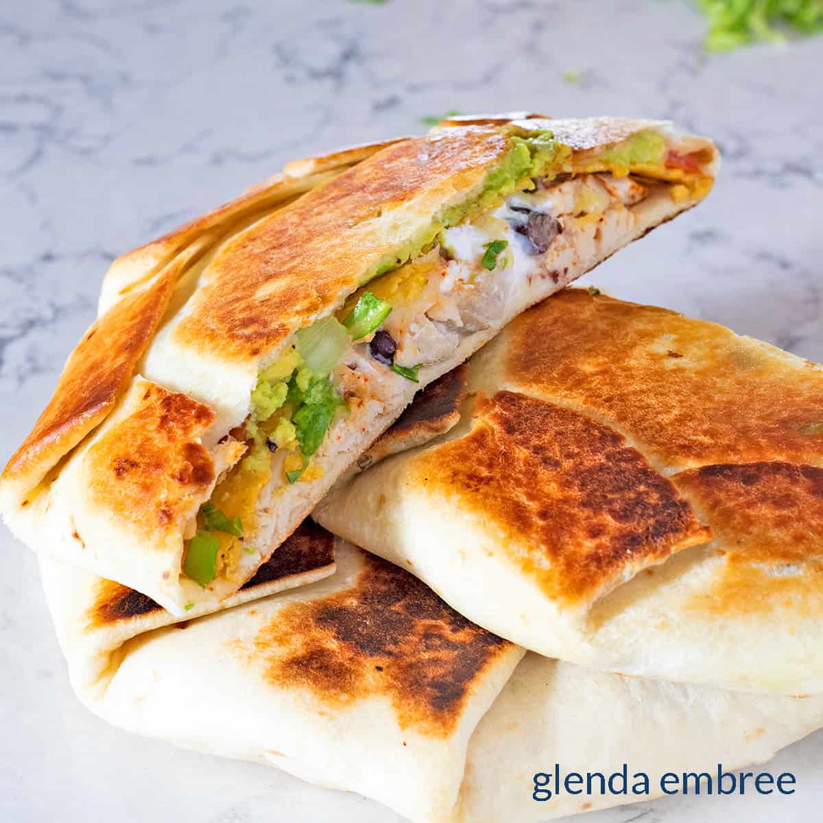 Chicken Crunch Wrap Supreme cut in half and placed on marble countertop - chicken, black beans, cheese, lettuce, tomato, sour cream and a crisp tostada shell wrapped in a burrito size flour tortilla to create a crisp, hand-held pocket sandwich.