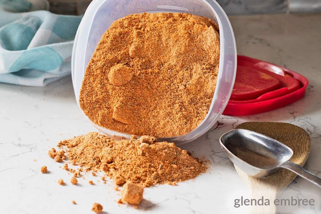 Homemade Cajun Seasoning spilling from a plastic container onto a marble countertop. Wooden spoon and measuring spoon on the counter.