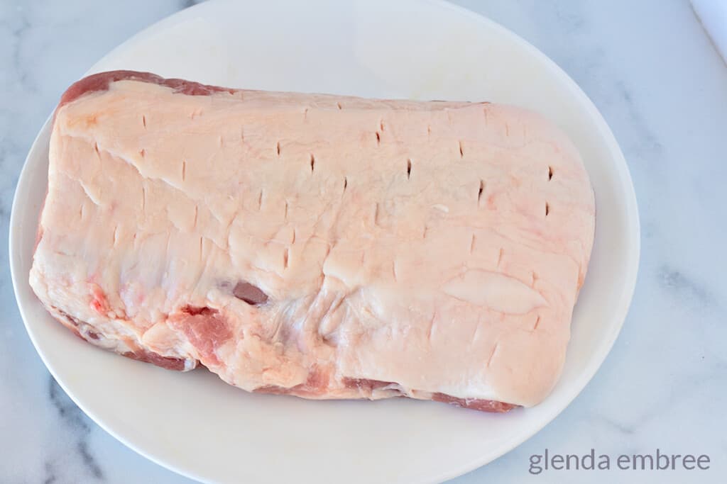 raw pork loin roast on a white plate. Roast has been pierced multiple times to allow seasonings to seep through the roast as it cooks.