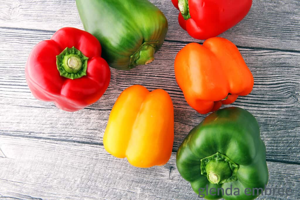 two red bell pepper, a yellow, an orange and twogreen bell peppers on a wooden table