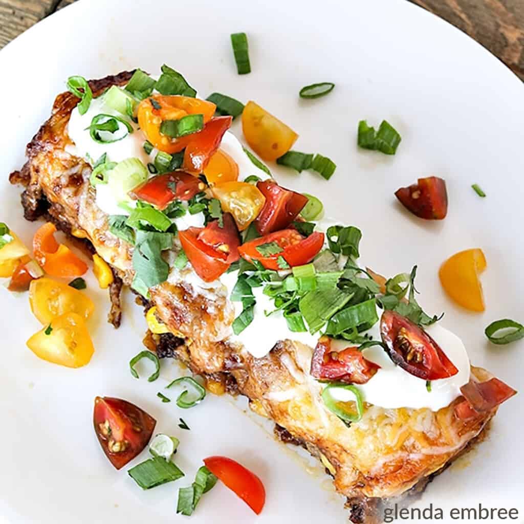Beef enchilada casserole garnished with sour cream, chopped tomatoes and green onions, served on a white plate - casserole recipes - 5 ingredient recipes