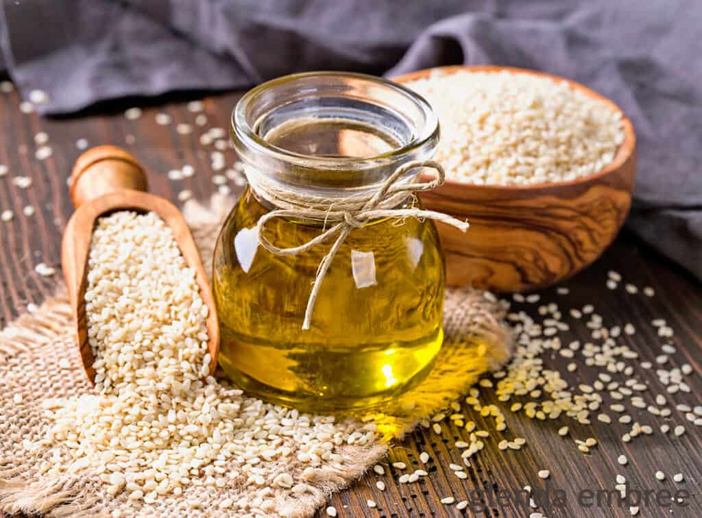 bottle of sesame oil on a wooden table with sesame seeds in a bowl and scoop as well as scattered on the table