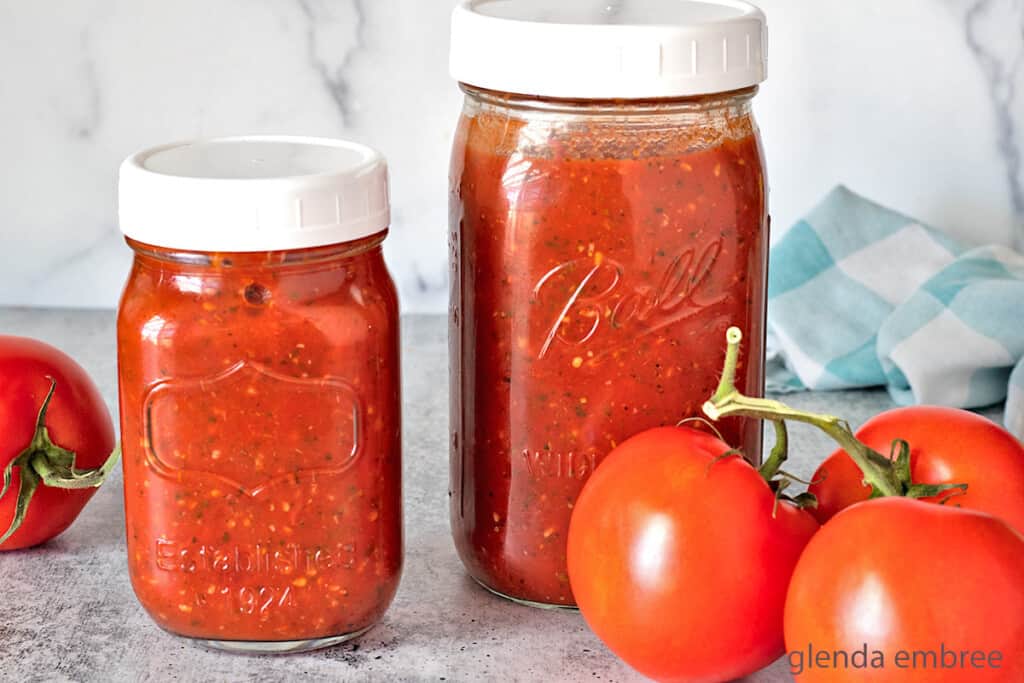 homemade marinara recipe in jars on concrete counter top with fresh tomatoes and a blue and white gingham napkin
