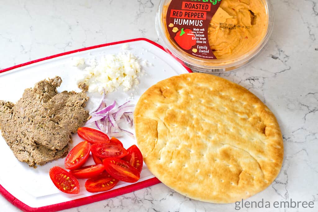 ingredients for a gyro pizza - pita flatbread, homemade gyro meat slices, halved cherry tomatoes, slivered red onion, feta cheese and roasted red pepper hummus.