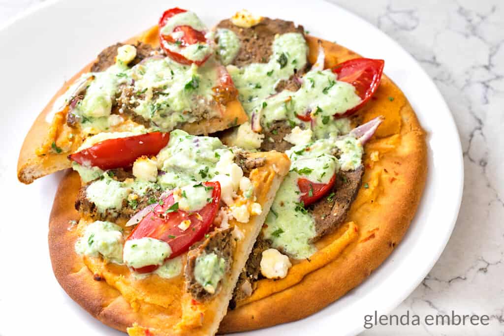 gyro pizza made on a pita flat bread with roasted red pepper hummus, homemade gyro meat, cherry tomatoes, slivered red onion, feta cheese, tzatziki sauce and a sprinkling of parsely
