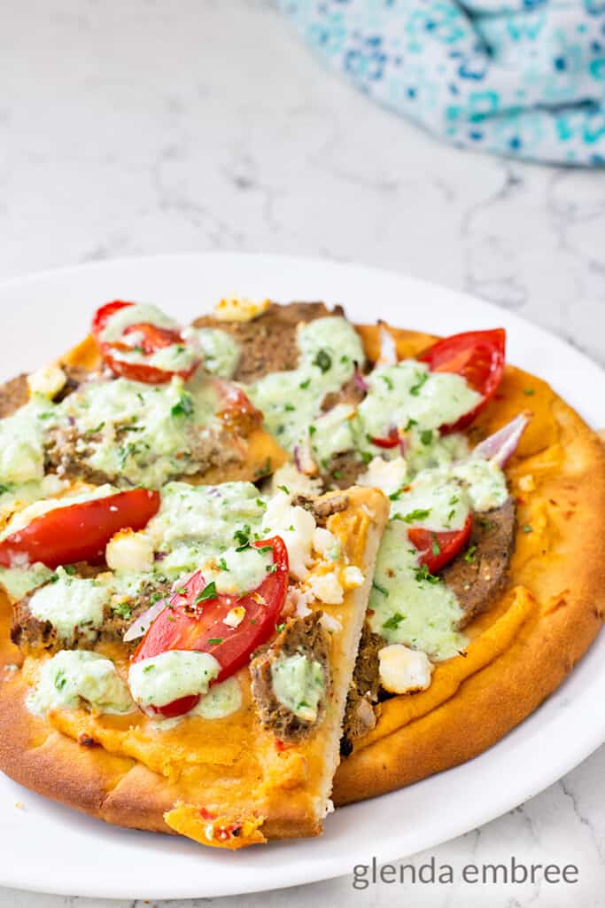 gyro pizza made on a pita flat bread with roasted red pepper hummus, homemade gyro meat, cherry tomatoes, slivered red onion, feta cheese, tzatziki sauce and a sprinkling of parsely