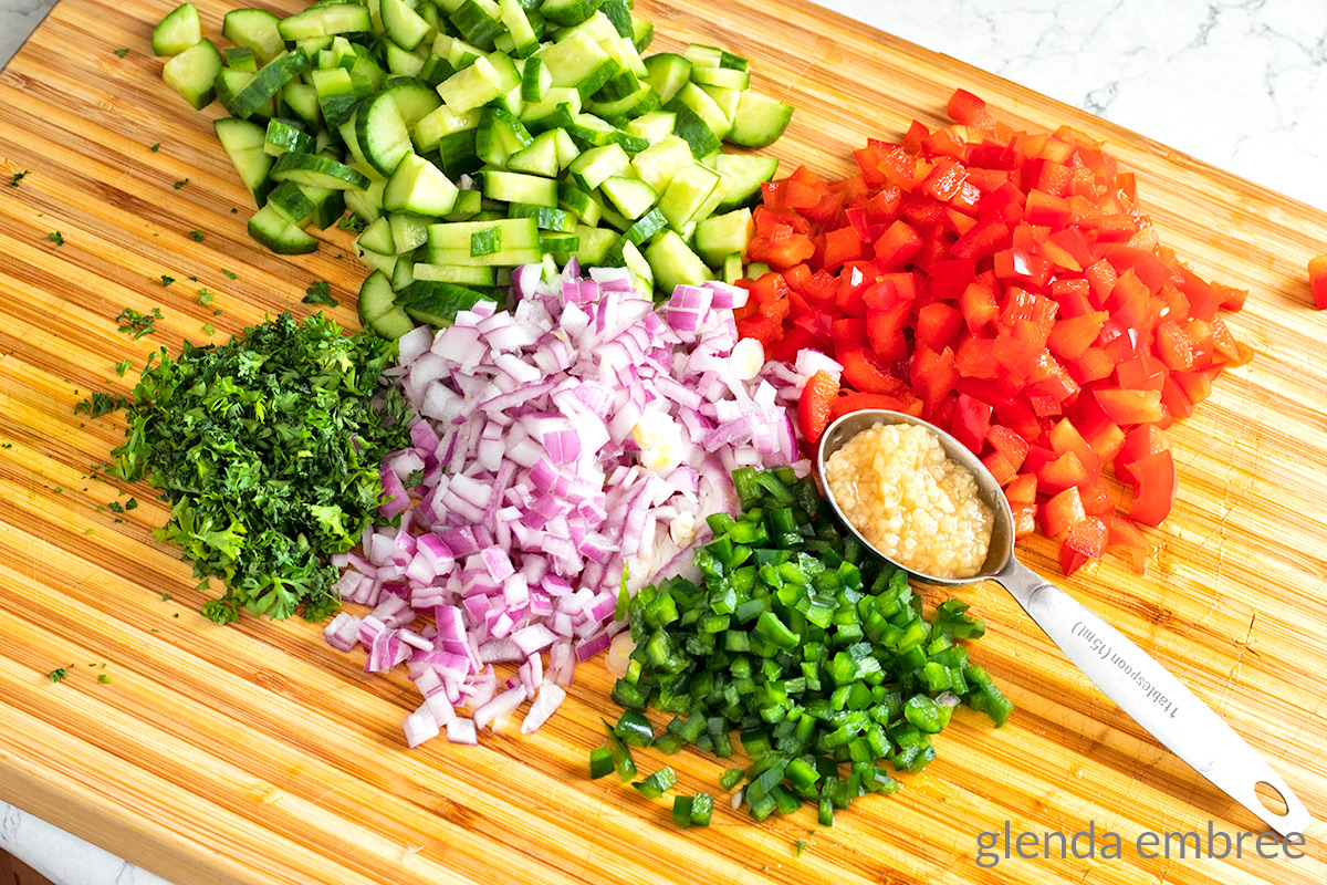 minced vegetables for pineapple salsa on a wooden cutting board - cucumber, red bell pepper, poblano pepper, red onion, garlic and parsley