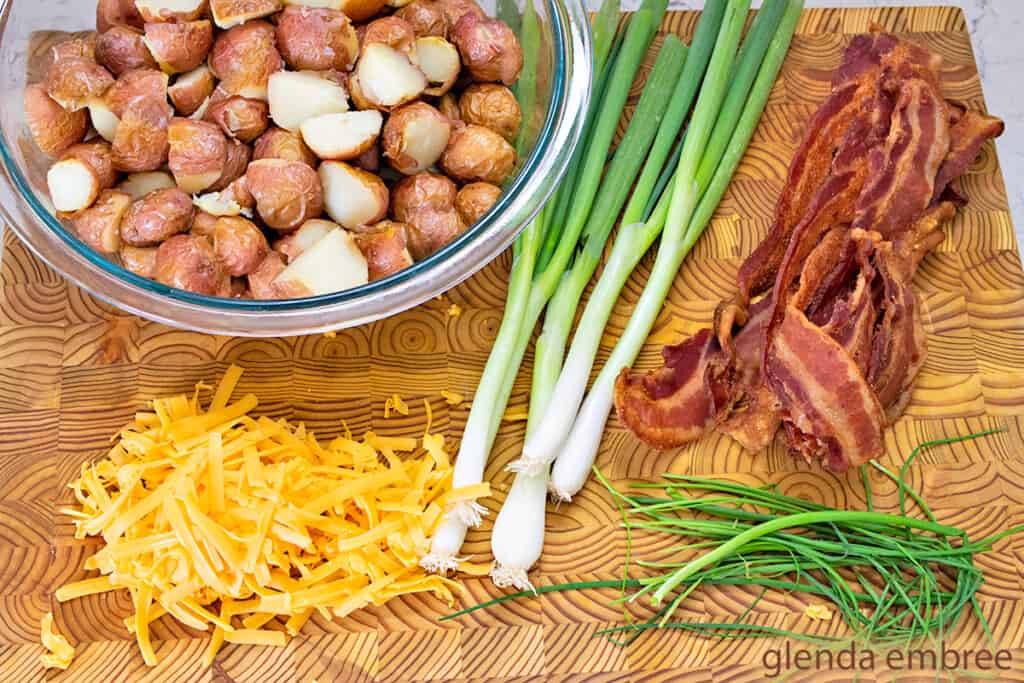 Roasted Red Potatoes, green onions, bacon, grated Cheddar cheese and whole chive stems on a wooden cutting board