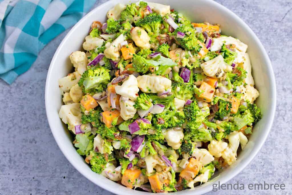 easy broccoli cauliflower salad recipe - salad in a round, white ceramic bowl ona concrete counter with a turquoise and white checked napkin