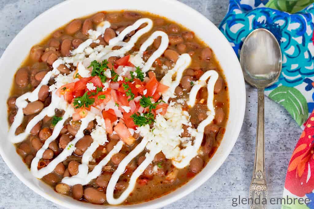 Slow Cooker Charro Beans in a white ceramic bowl and garnished with sour cream, diced tomatoes, green onion, parsley and queso fresco