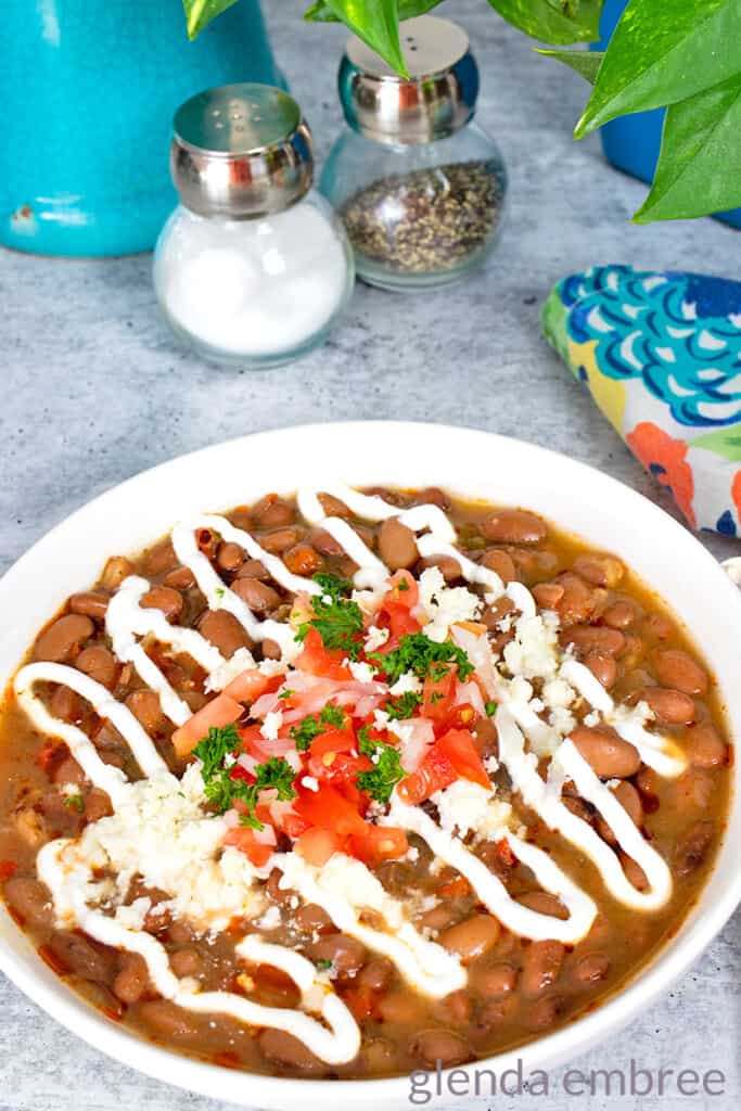 Slow Cooker Charro Beans in a white ceramic bowl and garnished with sour cream, diced tomatoes, green onion, parsley and queso fresco - pinto beans recipe