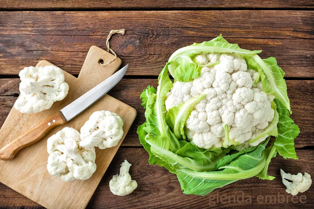 head of cauliflower on a wooden table next to a wooden cutting board with a knife and cauliflower florets on it