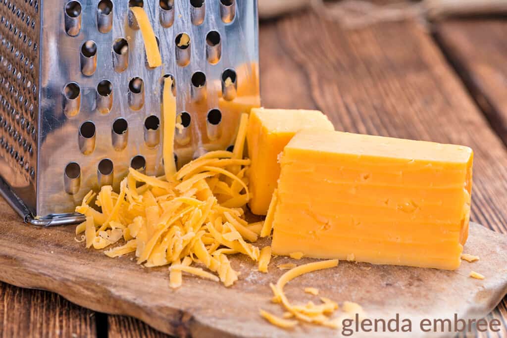 Grating cheddar cheese with a box grater.  Block of cheddar cheese and grater with a few shreds of grated cheese on a wooden cutting board.