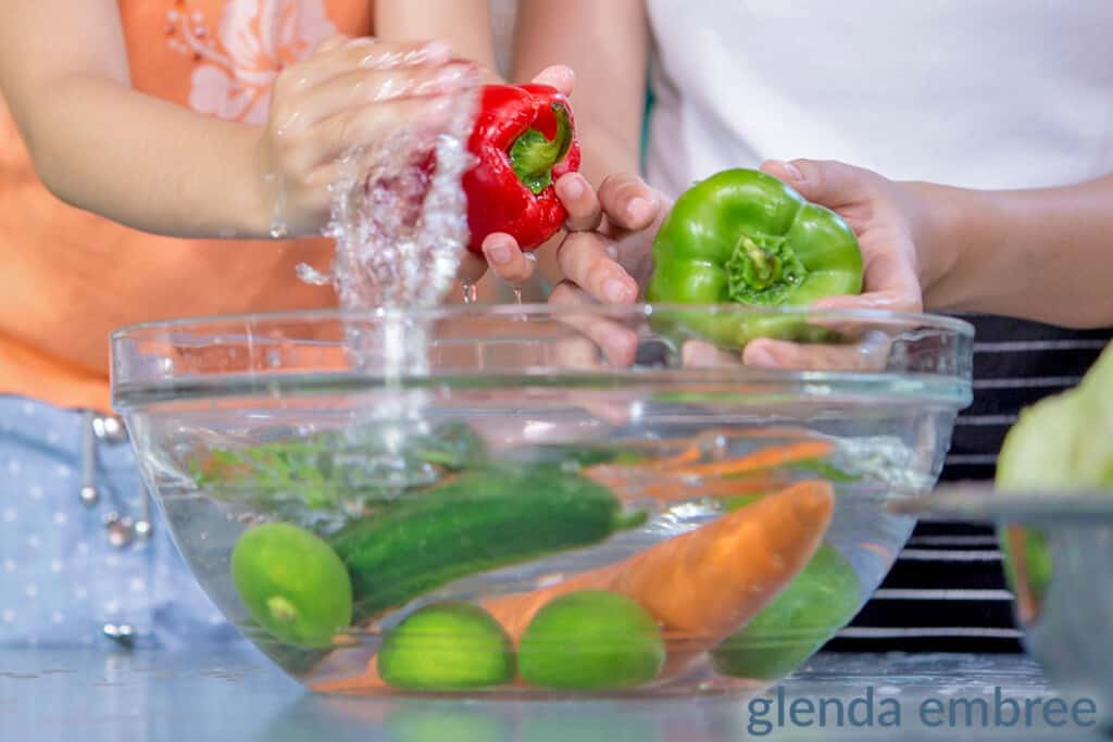 washing produce in a clear bowl.  Two sets of hands, ana adult and a child, washing peppers, carrots, cucumbers and limes.