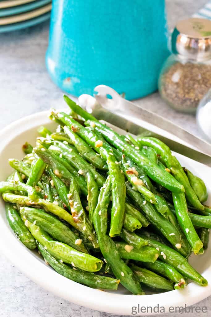 Air fryer green beans with garlic butter sauce served in an oval-shaped white bowl. The bowl is on a concrete counter which has a turquoise and white kitchen towel draped across.