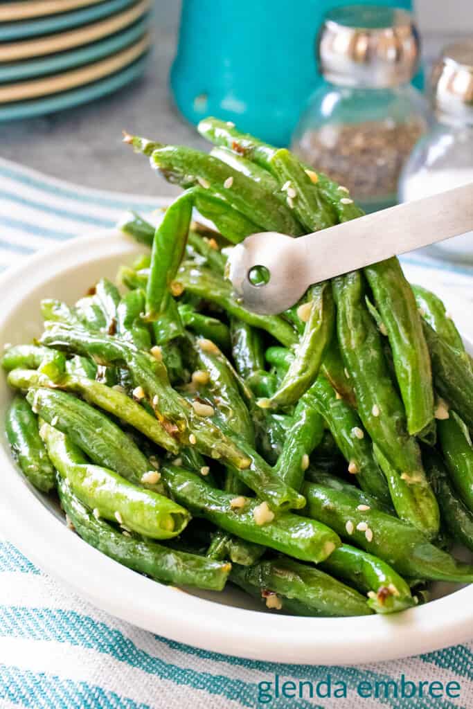 Air fryer green beans with garlic butter sauce served in an oval-shaped white bowl. The bowl is on a concrete counter which has a turquoise and white kitchen towel draped across. Green beans are being lifted from the bowl with metal tongs.