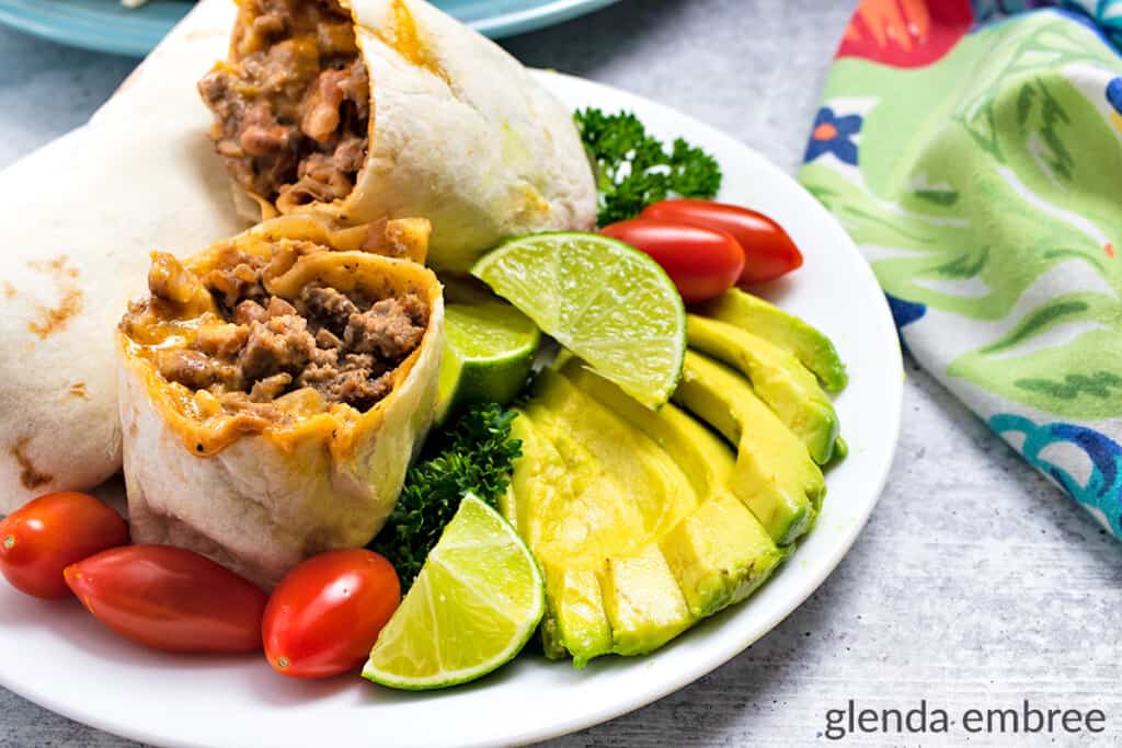 Beef and Bean Burritos on a white plate with sliced avocado, cherry tomatoes, parsley and lime wedges
