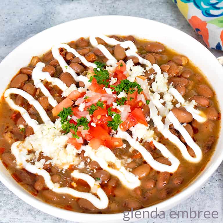 Slow Cooker Charro Beans in a white ceramic bowl and garnished with sour cream, diced tomatoes, green onion, parsley and queso fresco