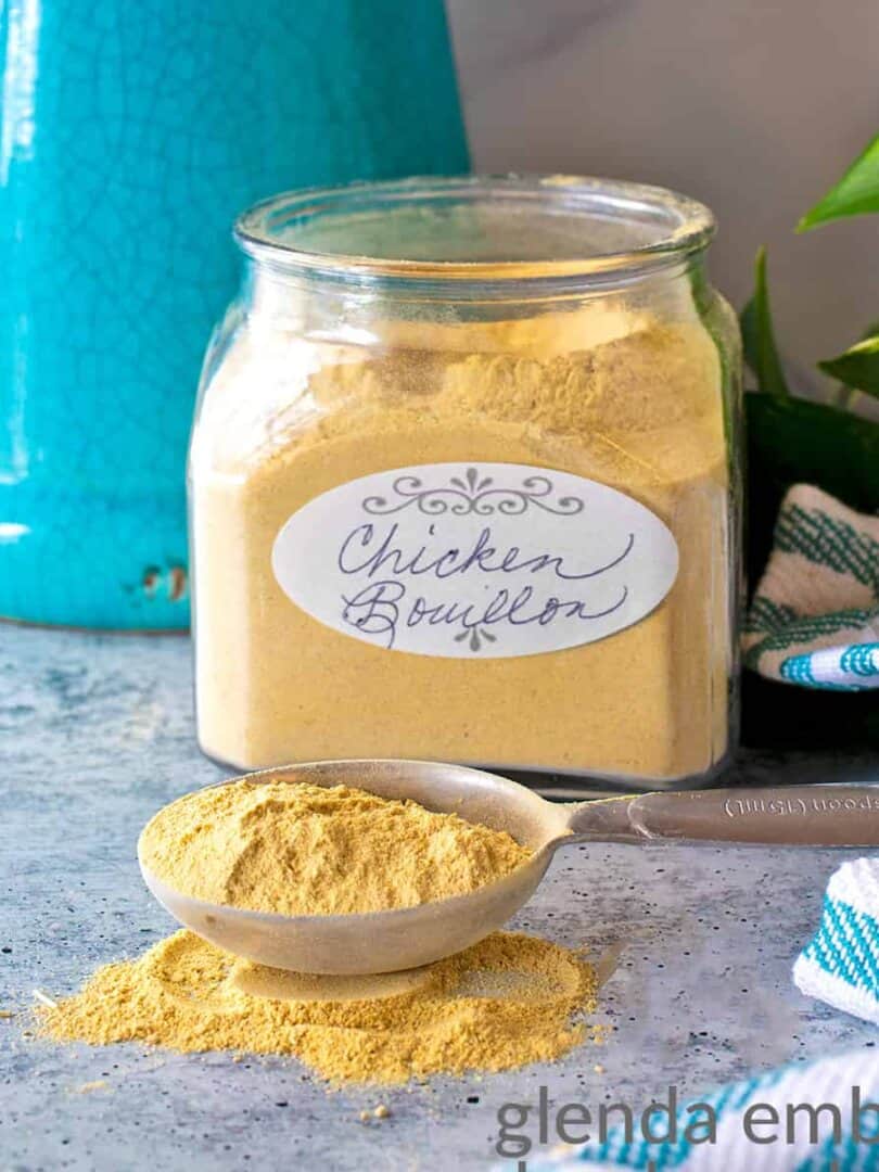 chicken bouillon powder in an apothecary jar with a Tablespoon of chicken stock powder spilling out onto the concrete counter top. There is a turquoise stoneware pitcher, a philedendron and and turqupise and white striped kitchen towel in the background