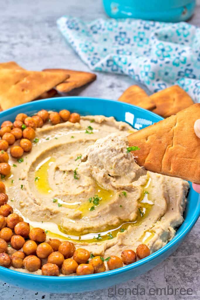 Creamy Hummus with Roasted Garlic in a blue stoneware bowl. Garnished with cripy chickpeas, olive oil drizzle and served with pita chips