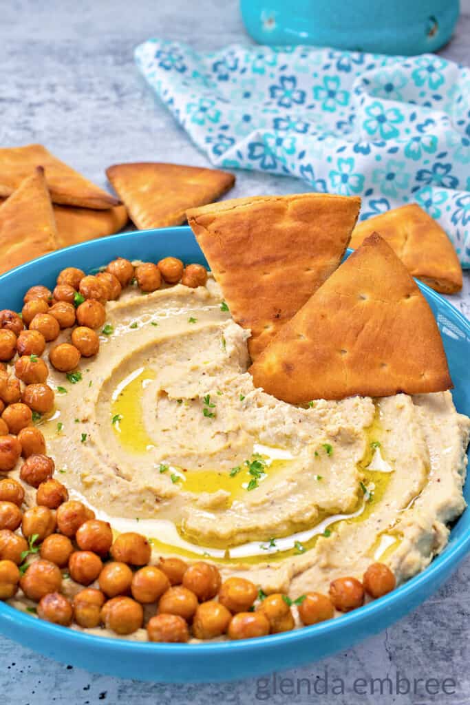 Creamy Hummus with Roasted Garlic in a blue stoneware bowl. Garnished with cripy chickpeas, olive oil drizzle and served with pita chips