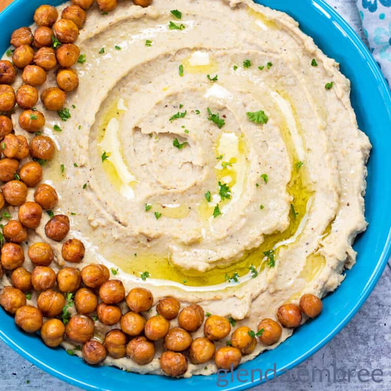 creamy hummus in a blue stoneware bowl with pita chips and a blue print fabric napkin on a concrete countertop