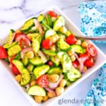 Mediterranean Cucumber Tomato Salad in a square serving bowl next to a blue and white print napkin. Potted plant and salt shaker in the background.