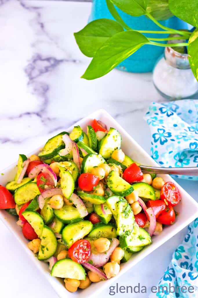 Mediterranean Cucumber Tomato Salad in a square serving bowl next to a blue and white print napkin. Potted plant and salt shaker in the background.
