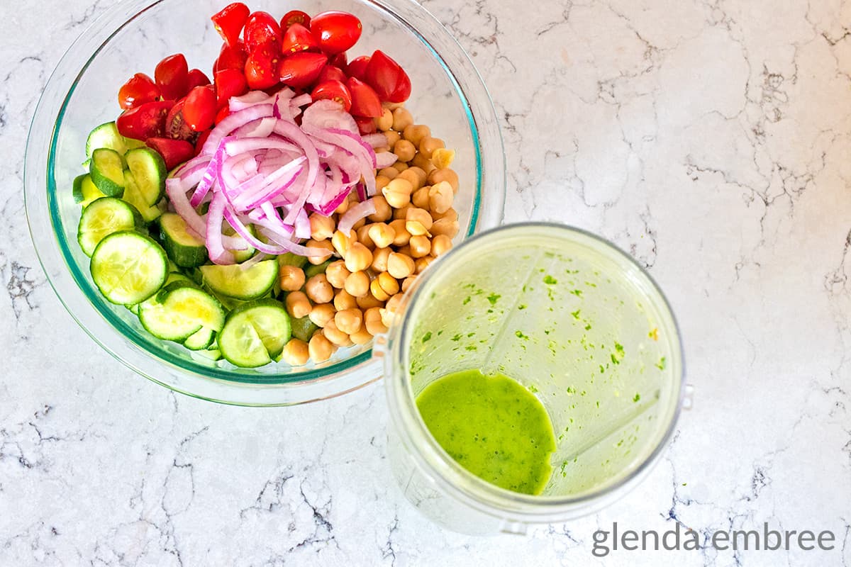 chopped tomatoes, red onion slivers, chickpeas, sliced cucumber in a clear glass mixing bowl next to a blender pitcher with homemade dressing