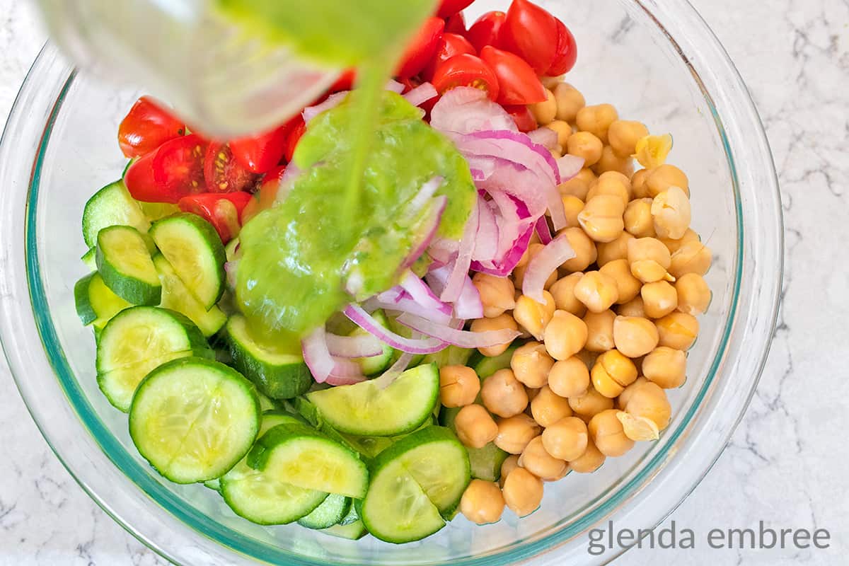 dressing being poured over chopped tomatoes, red onion slivers, chickpeas, sliced cucumber in a clear glass mixing bowl