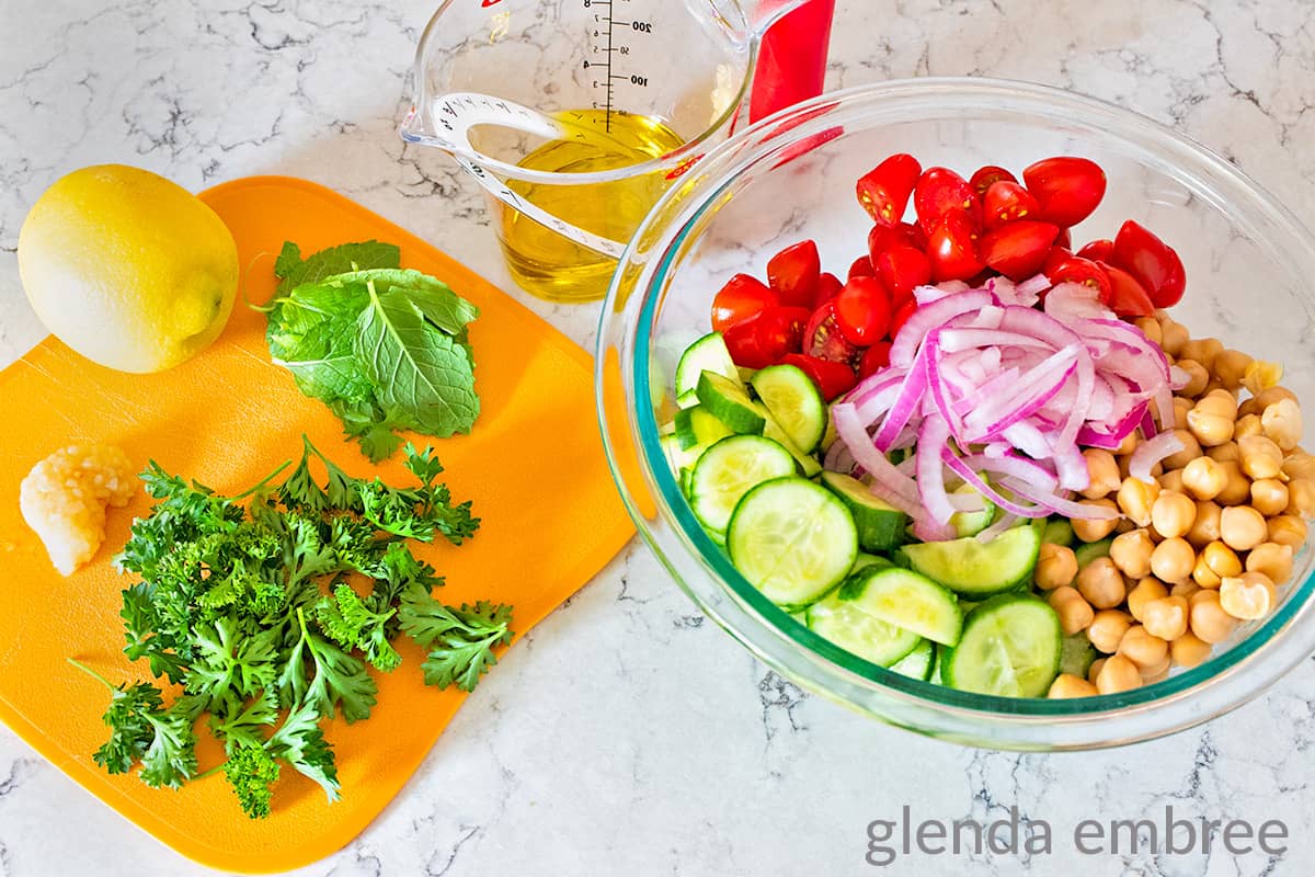chopped tomatoes, red onion slivers, chickpeas, sliced cucumber in a clear glass mixing bowl next to a cutting board with a lemon, fresh mint leaves, fresh parsely and garlic. Liquid measuring cup with olive oil in the background.