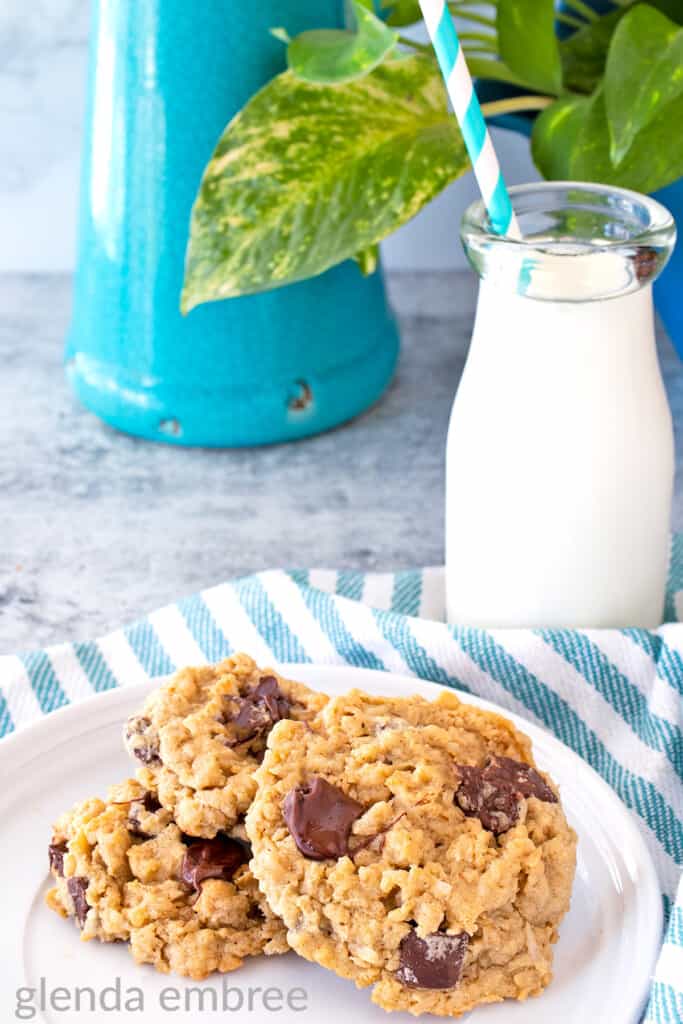 Ranger Cookies on a white plate sitting on a concrete counter with a bottle of milk next to a turquoise vase