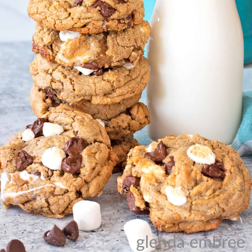 smores cookies stacked next to a milk bottle with a blue and white striped straw. Two cookies are laying on the counter in front of the cookie stack and milk bottle with marshmallows and chocolate chips strewn around.