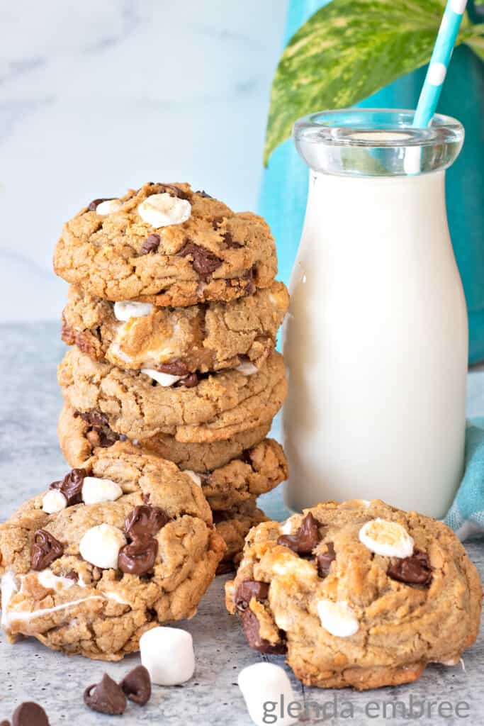 smores cookies stacked next to a milk bottle with a blue and white striped straw. Two cookies are laying on the counter in front of the cookie stack and milk bottle with marshmallows and chocolate chips strewn around.